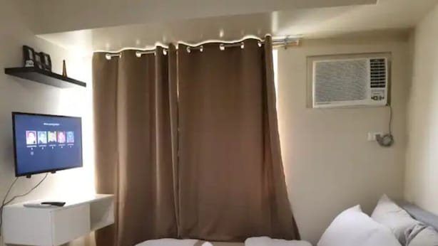 Hotellihuone, jossa on sänky, jalustalla oleva televisio, ikkuna verhoilla ja seinälle asennettu ilmastointilaite.