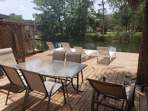 Relax on the shaded deck with a glass-top table and wicker chairs, surrounded by lush greenery and a serene water view.