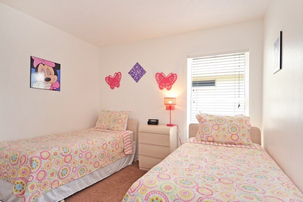 A bedroom with two single beds, a nightstand, a lamp, and wall decorations including a picture of a cartoon character and butterfly-shaped ornaments.