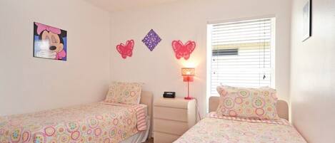 A bedroom with two single beds, a nightstand, a lamp, and wall decorations including a picture of a cartoon character and butterfly-shaped ornaments.