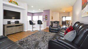 A living room with a flat-screen TV, a wooden entertainment unit, a black leather sofa, and a gray shag rug. - Home from home! Updated - private pool & hot tub - gated resort (Clermont)