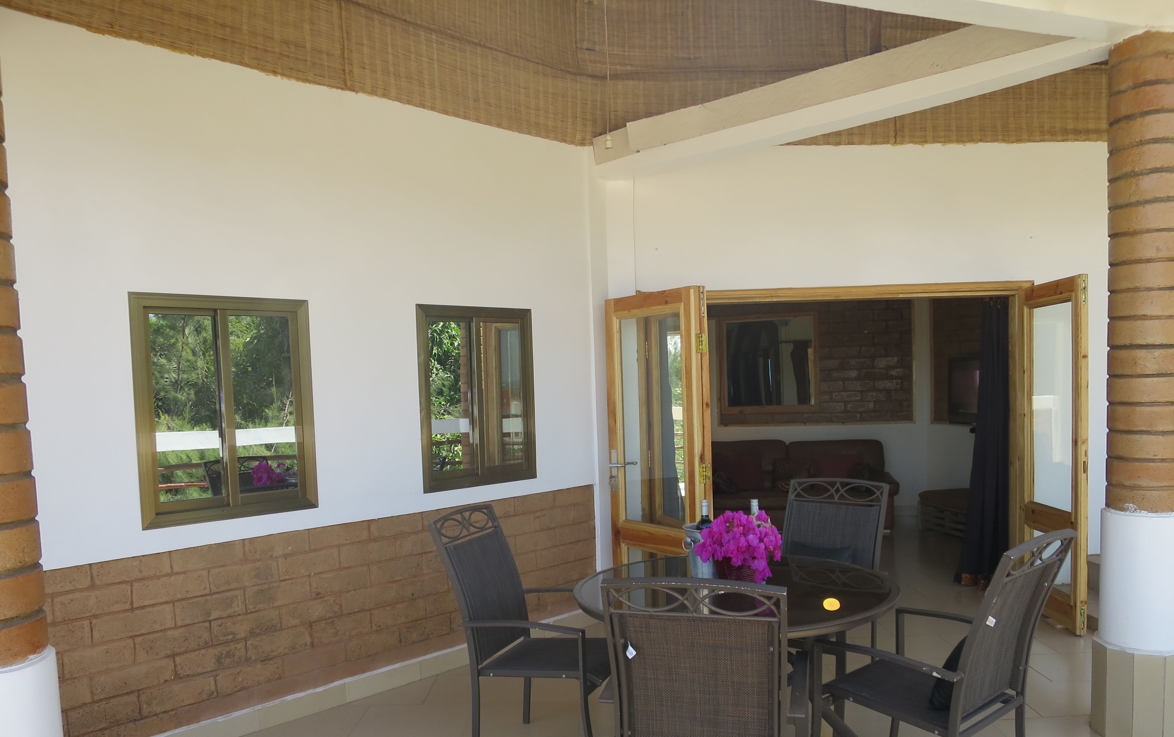 Uma área de estar coberta ao ar livre, com mesa e cadeiras, porta de vidro que dá para um espaço interno, e parede de tijolos.