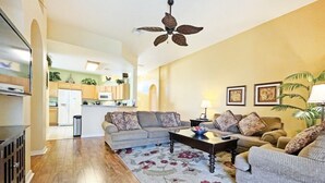 A living room with a sofa, a coffee table, and a patterned rug. There is a ceiling fan, a kitchen in the background, and a trash can. - Superior family vacation home (Haines City)