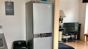 A compact kitchen with a stainless steel refrigerator, a trash bin, a dining area with a TV, and a small table.