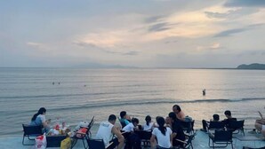Eine Gruppe von Menschen sitzt an Tischen auf einer Terrasse mit Blick auf ein ruhiges Meer und einen Sonnenuntergang im Hintergrund.