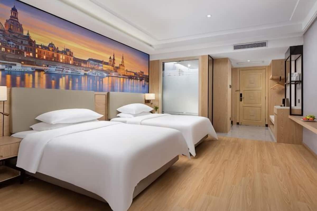 Une chambre d’hôtel avec deux lits, une fresque représentant un paysage urbain, un plancher en bois, un bureau avec un miroir et une porte donnant sur une autre pièce.
