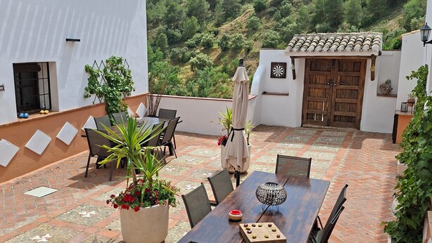 A patio with a wooden table and chairs, surrounded by potted plants and a view of a hillside. - Casa Los Molinos with heated pool in the middle of nature (Montefrio)