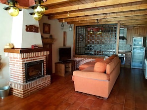 A living room with a brick fireplace, a sofa, a television, and a kitchen area visible through a glass partition.