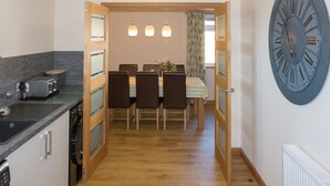 A compact kitchen with modern appliances, a dining area with a wooden table and chairs, and a large wall clock. - Bright modern apartment (Keiss)