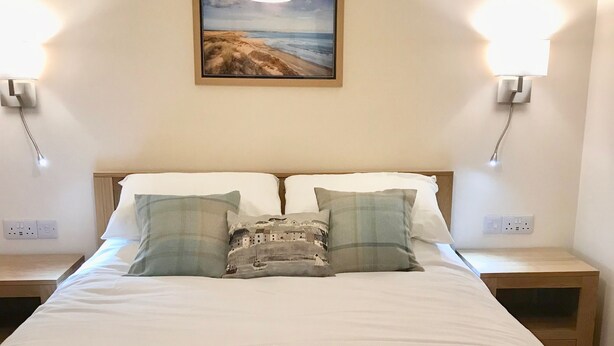 A neatly made bed with a folded towel in the center, two bedside lamps, a wall-mounted painting, and a nightstand with a power outlet. - Bright modern apartment (Keiss)