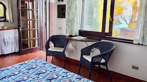 Ein Zimmer mit einem Bett, zwei Korbstühlen, einem kleinen Tisch, einem Fenster mit Vorhängen und einer Tür mit Spiegel.