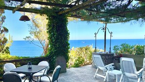 Eine Terrasse mit Sitzgelegenheiten im Freien, einem Steintisch und Blick aufs Meer.