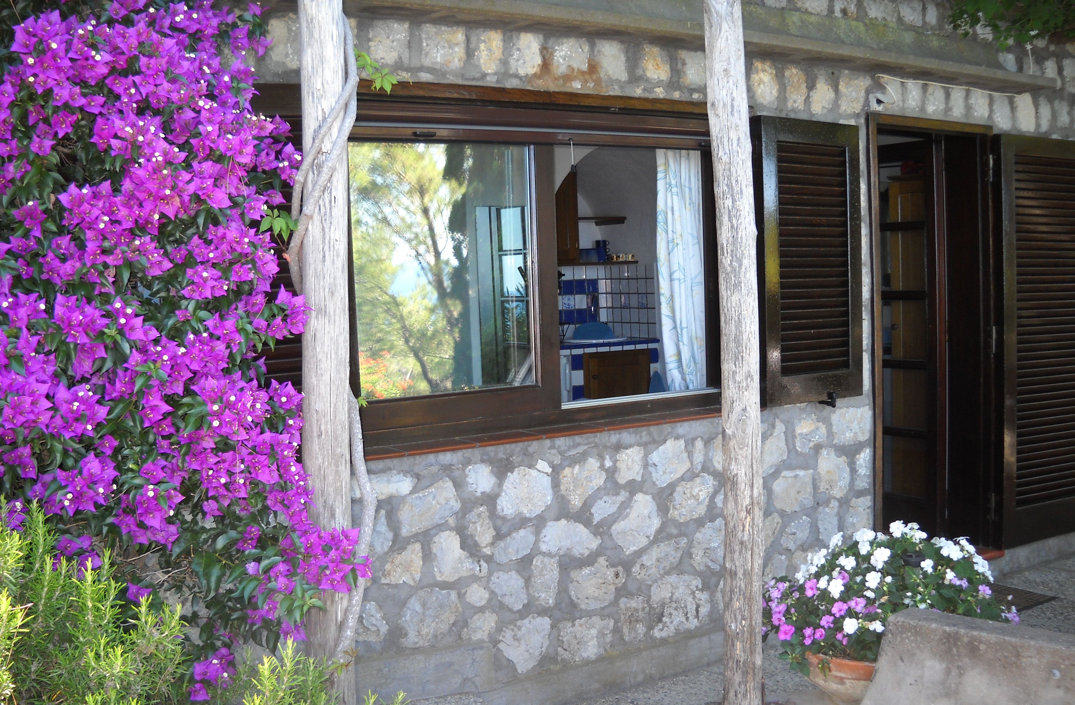 Ein Steingebäude mit Fenster und Fensterläden, umgeben von violettem Bougainvillea und einem Blumentopf mit weißen Blüten.