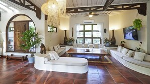 A spacious living area with a large sofa, a coffee table, and a dining table. There are several potted plants and a television mounted on the wall. - Paradise Beach Lodge (Cóbano)