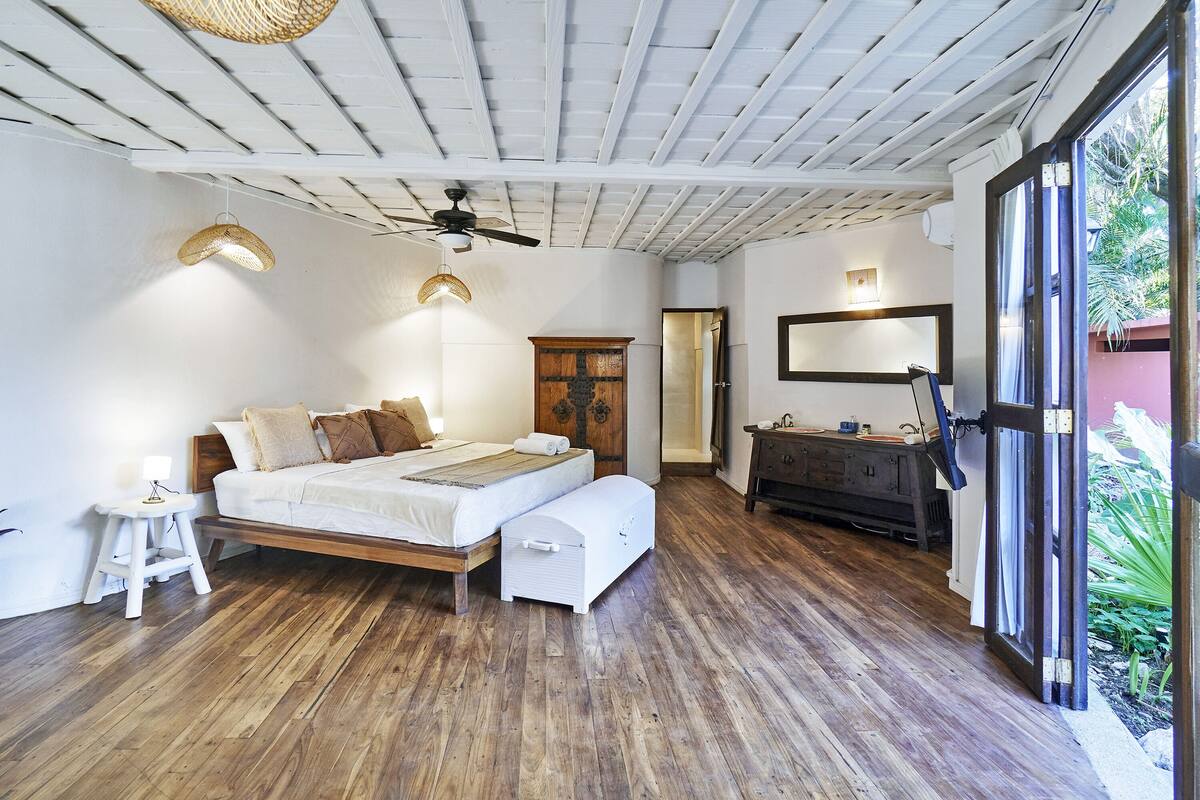 A bedroom with a wooden bed, a white bench, a dresser, a ceiling fan, and a large window with a view of greenery.