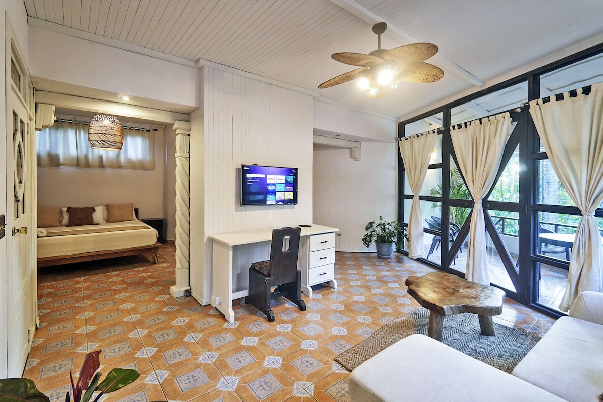 A room with a bed, desk, and seating area, featuring a large window with curtains and a ceiling fan.