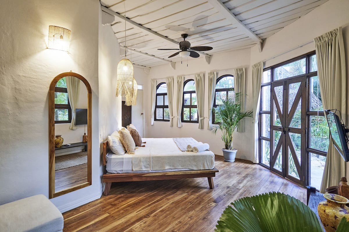 A bedroom with a wooden bed, large windows, a ceiling fan, and a potted plant.