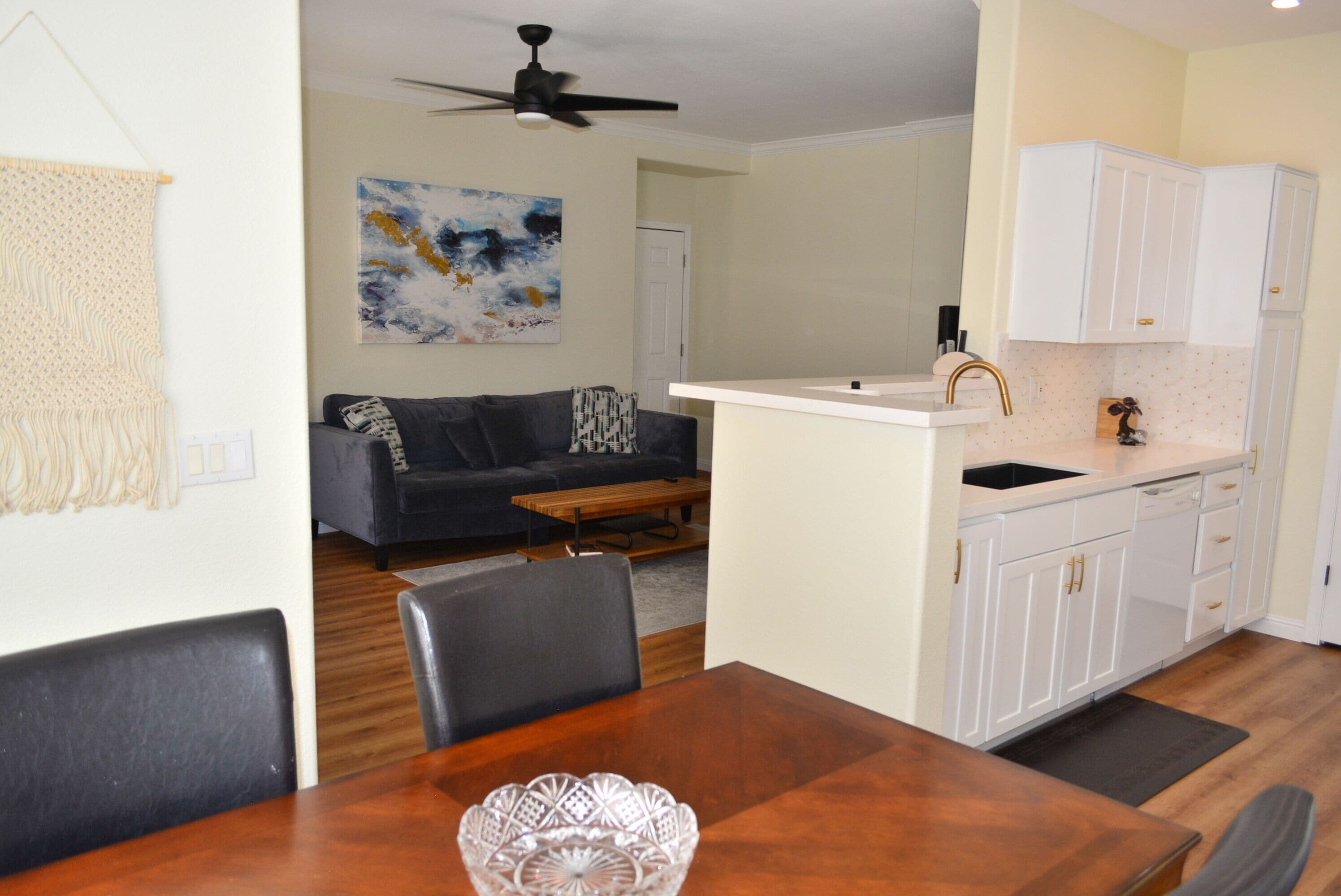 A modern kitchen with white cabinets and a black faucet, a dining area with a wooden table and chairs, and a living room with a gray sofa and a coffee table.
