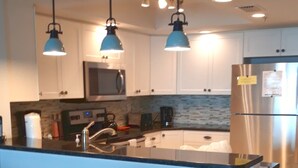A modern kitchen with white cabinets, a stainless steel refrigerator, a central island with bar stools, and pendant lighting. - DIRECT BEACHFRONT - PENTHOUSE CORNER UNIT!!  Totally Renovated! (Indian Shores)