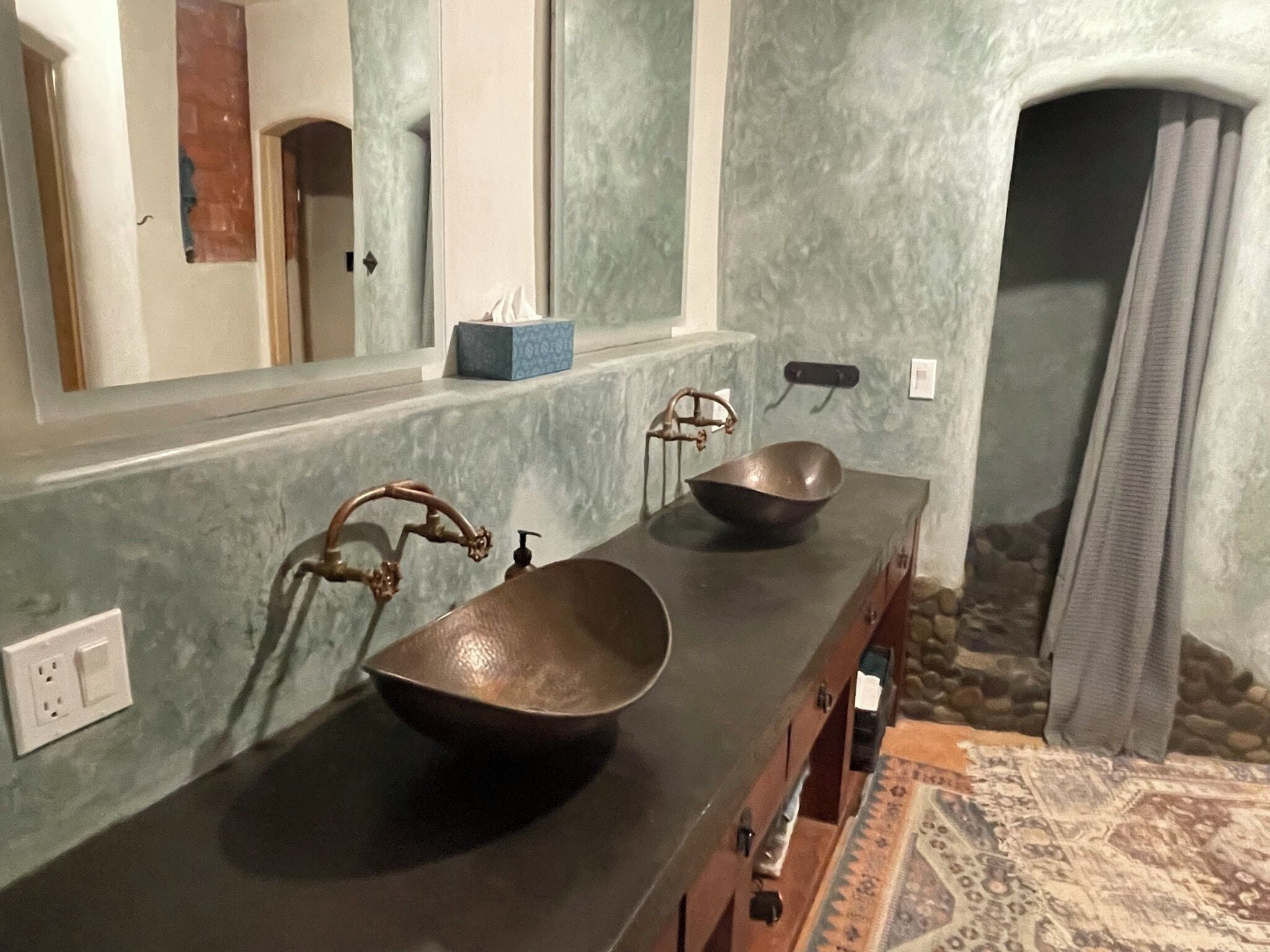 Casa de banho com um espelho grande, duas pias com torneira única, um porta-toalhas e uma parede de pedra.