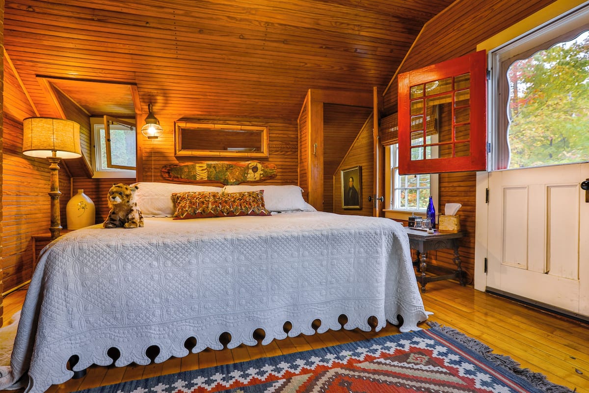 A cozy bedroom with a wooden ceiling, a large bed with a white quilt, a patterned rug, and a window with red shutters.
