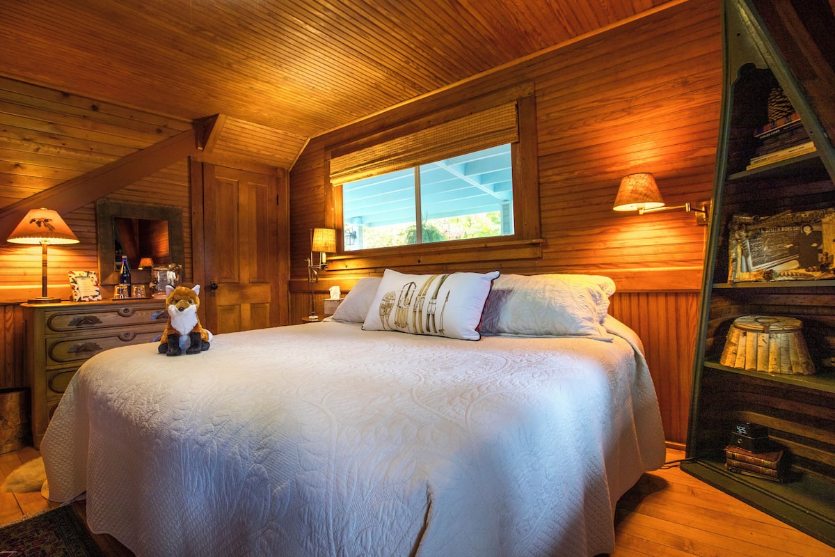 A cozy bedroom with a large bed, wooden walls, a window, and a nightstand with a lamp.