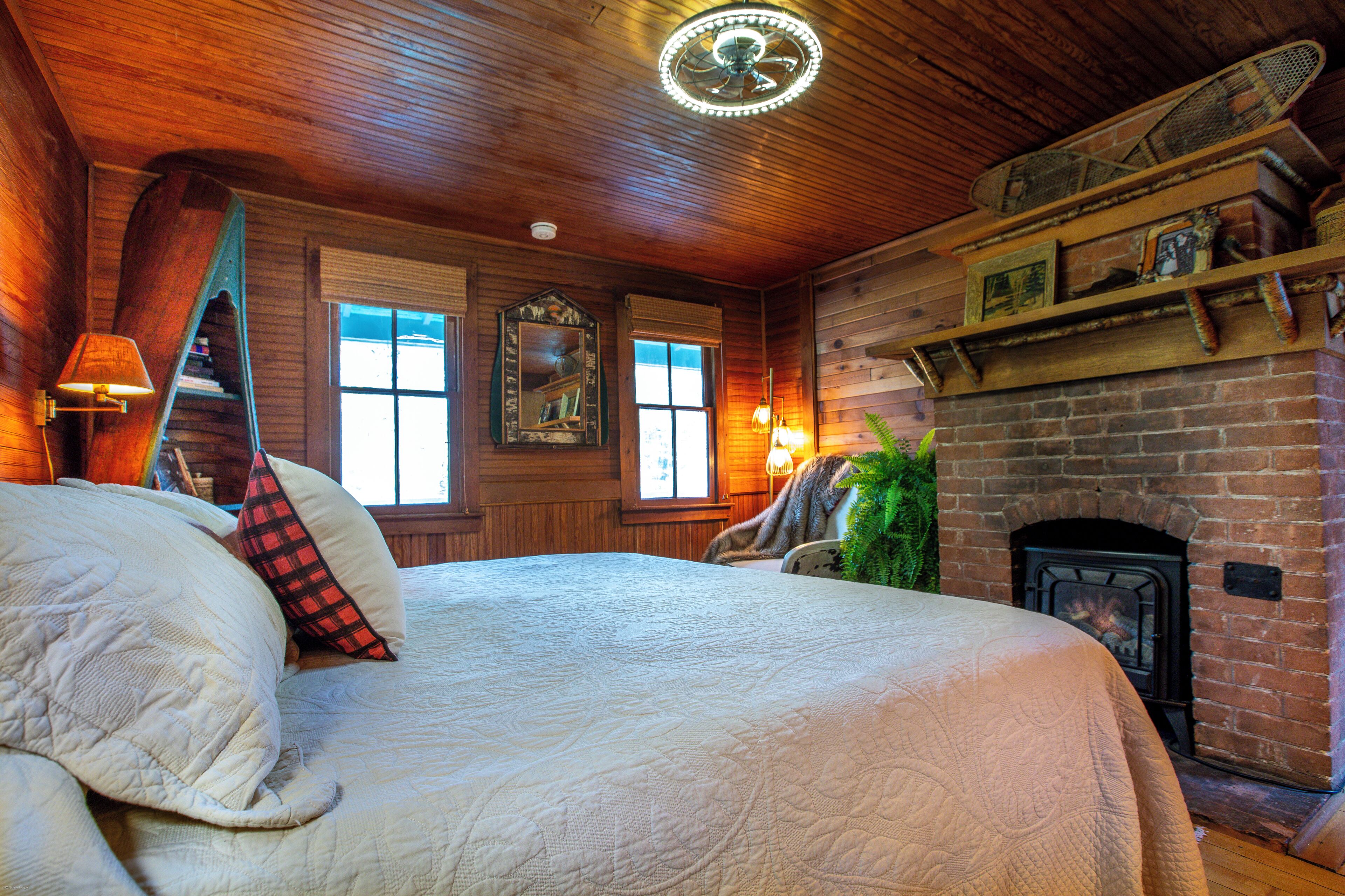 A room with a large bed, a fireplace, wooden walls, and a ceiling fan.