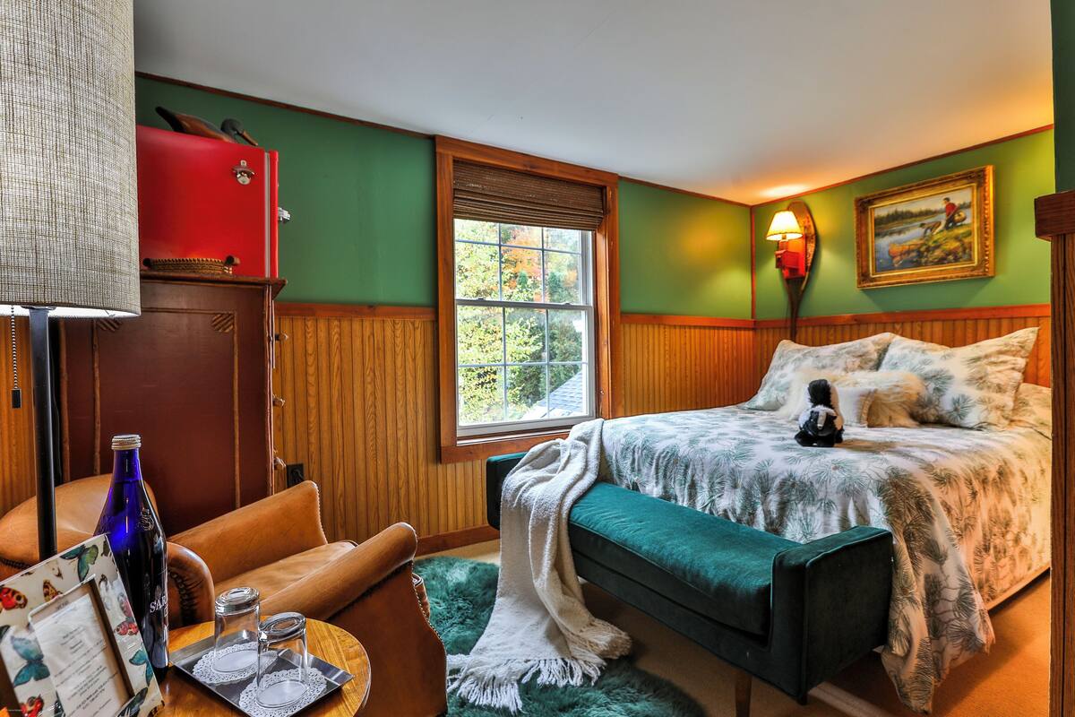 A room with a bed, a desk with a lamp, a window, and a painting on the wall.