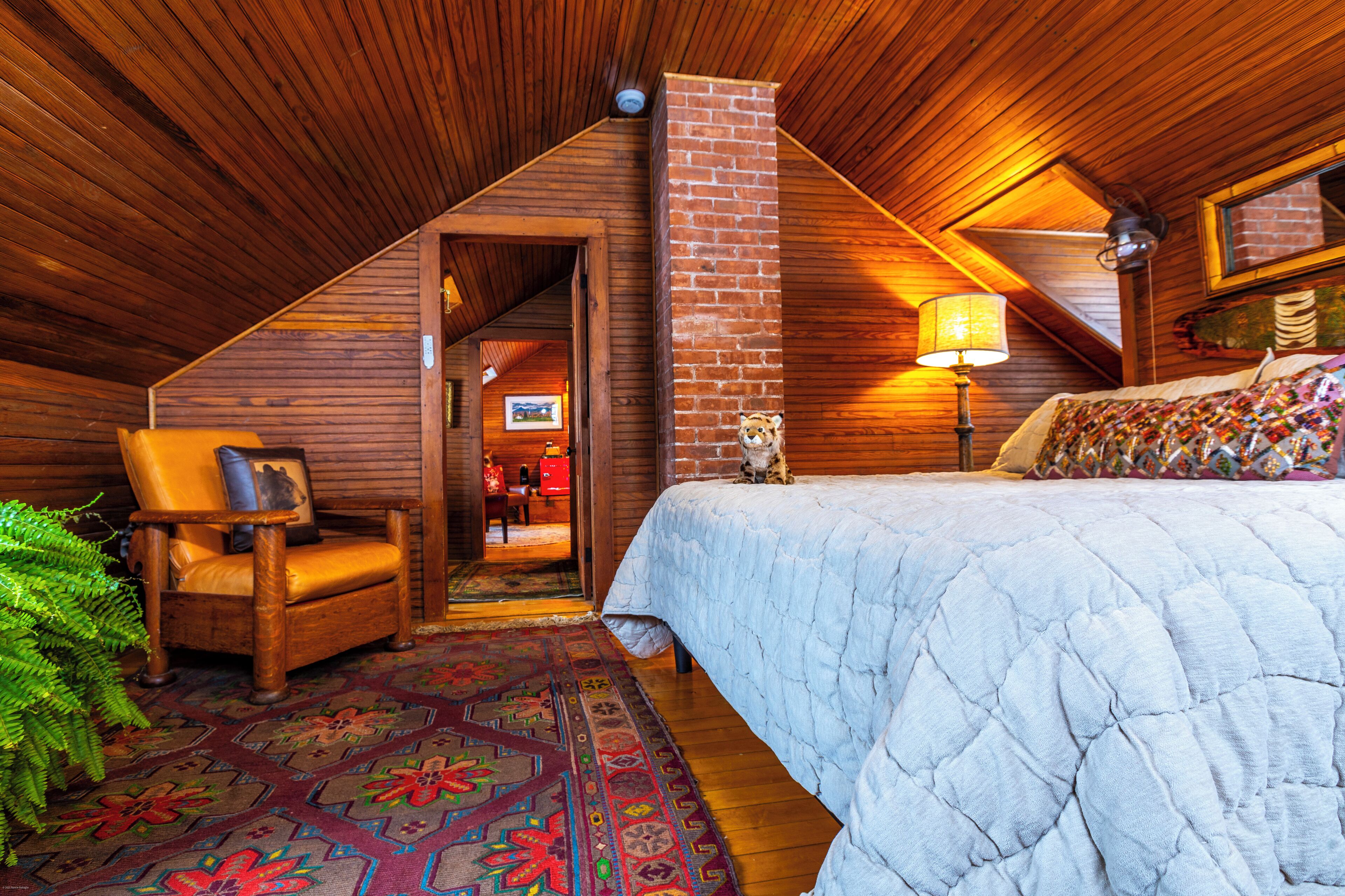 A cozy attic bedroom with a wooden ceiling, a brick chimney, a bed with a quilt, a chair with a cushion, and a patterned rug.