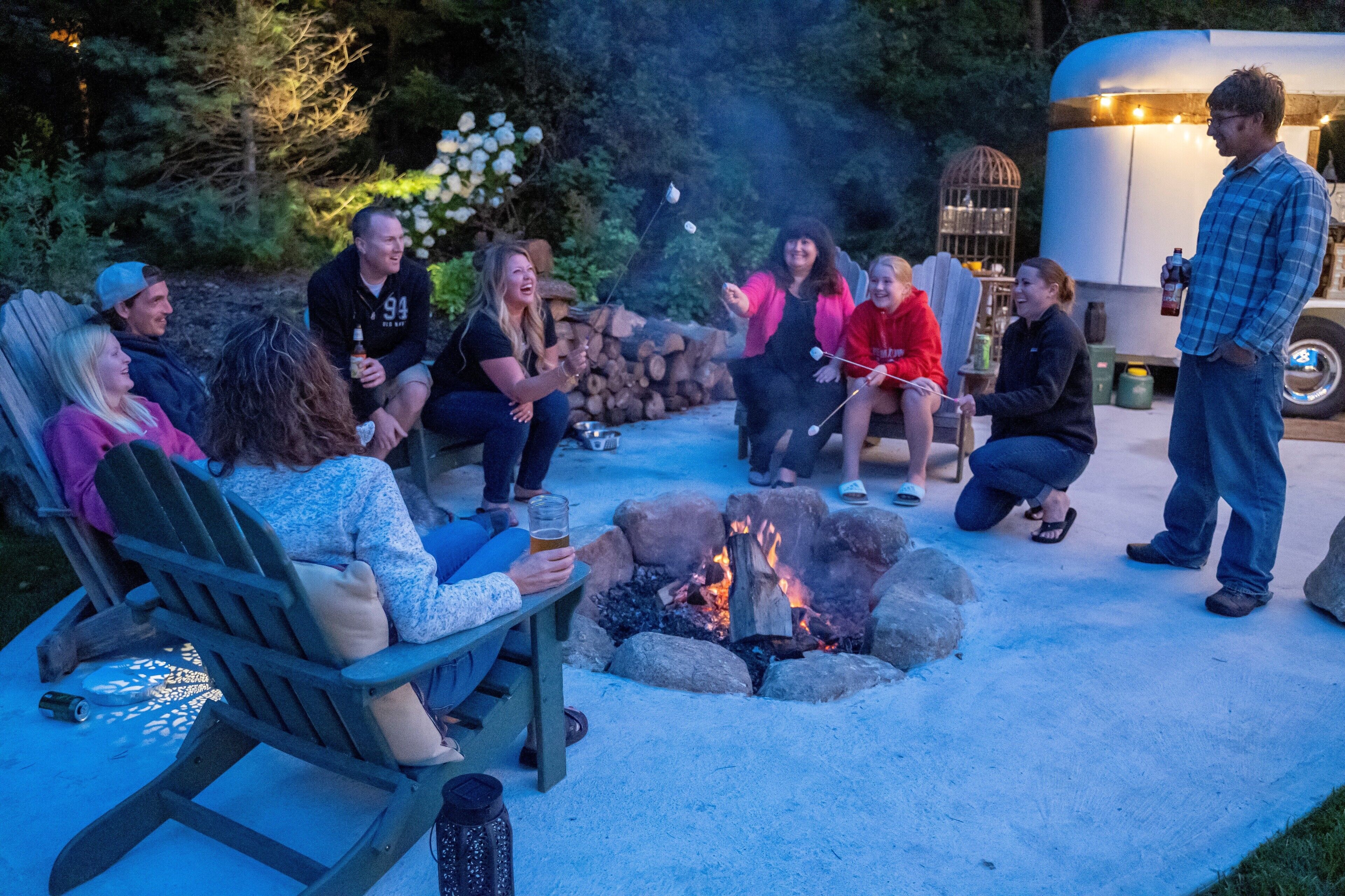 A group of people gathered around a fire pit in an outdoor setting with a trailer in the background.