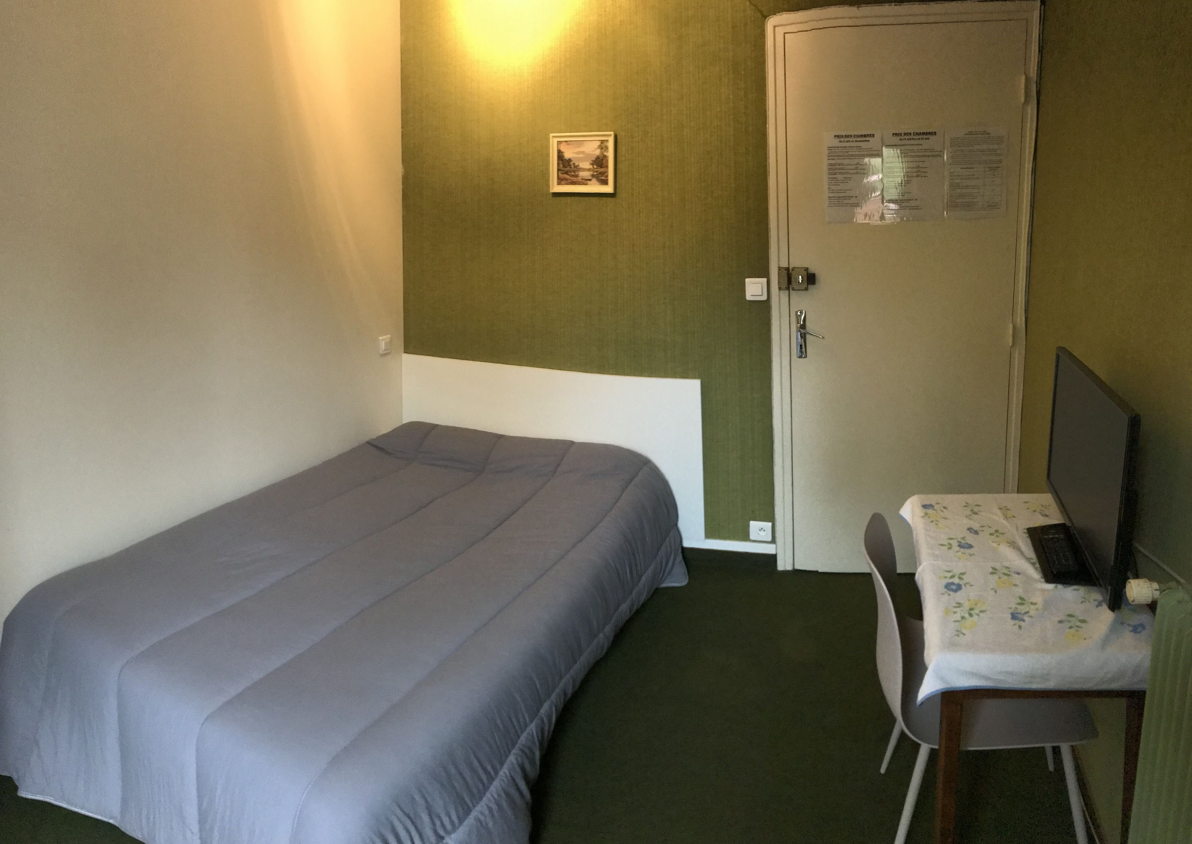 A small, single-bed room with a desk and chair, a door with a notice, and a framed picture on the wall.