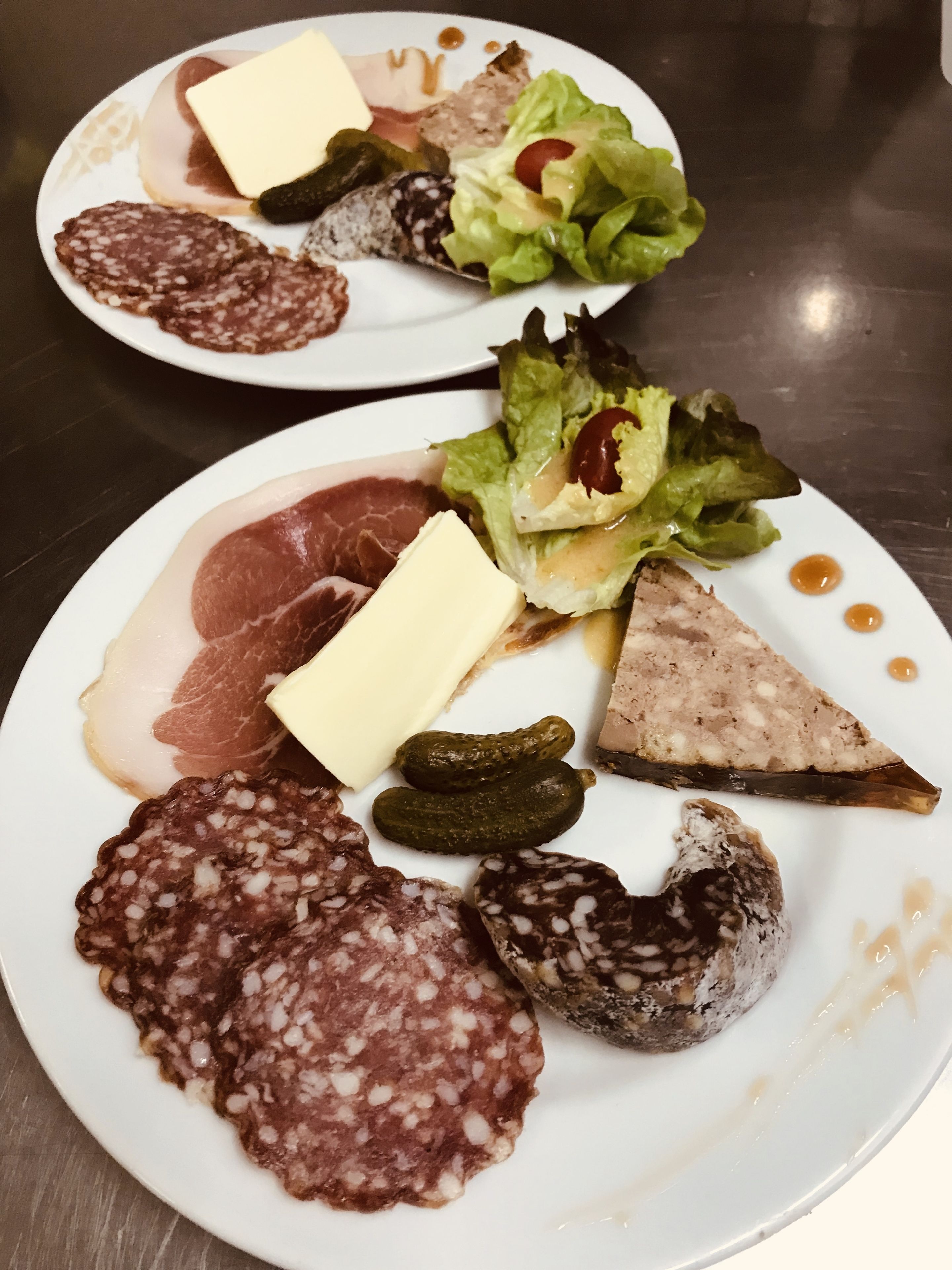 Two plates of assorted cured meats, cheese, and pickles on a stainless steel table.