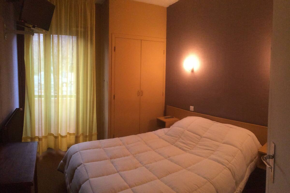 A hotel room with a bed, a desk, a wardrobe, and a wall-mounted light.