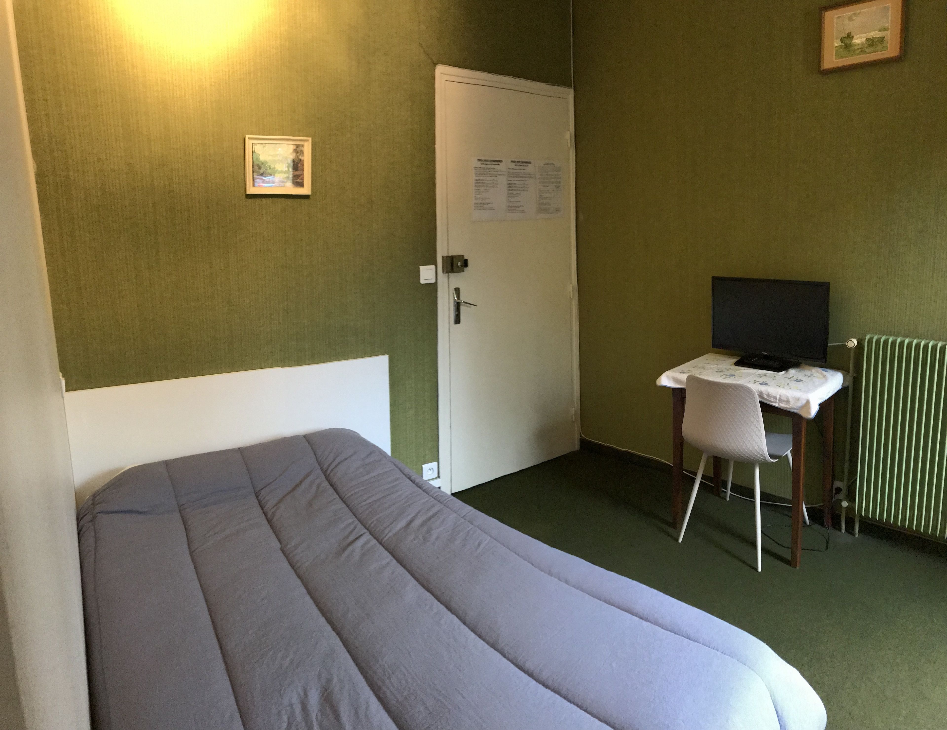 A hotel room with a bed, a desk with a computer, a chair, a door, and a radiator.
