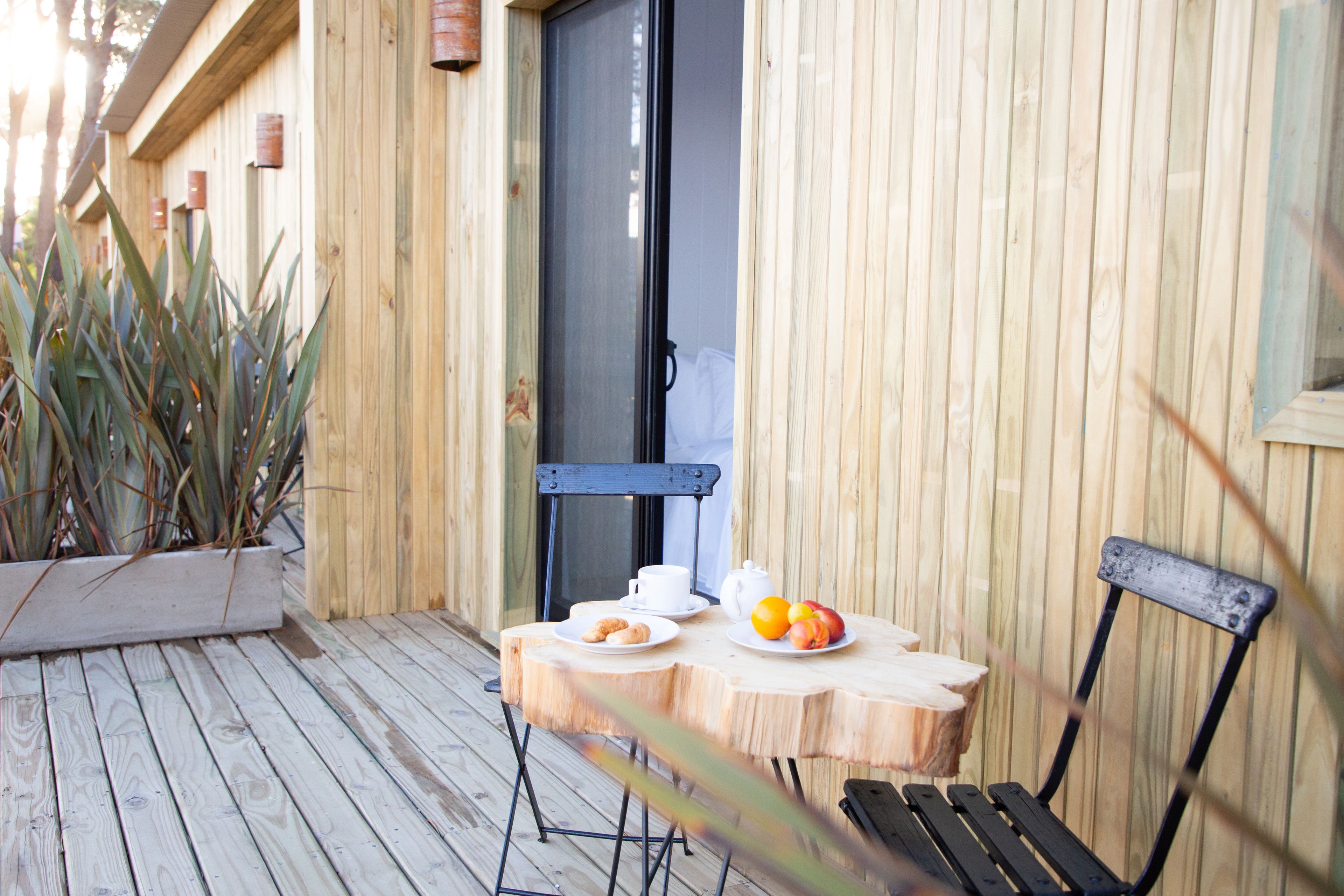 Una terraza de madera con una mesa lista para el desayuno, que incluye un plato de fruta y una taza.