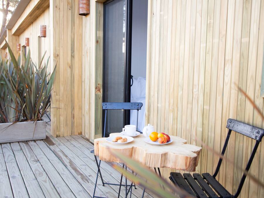 Una terraza de madera con una mesa lista para el desayuno, que incluye un plato de fruta y una taza.