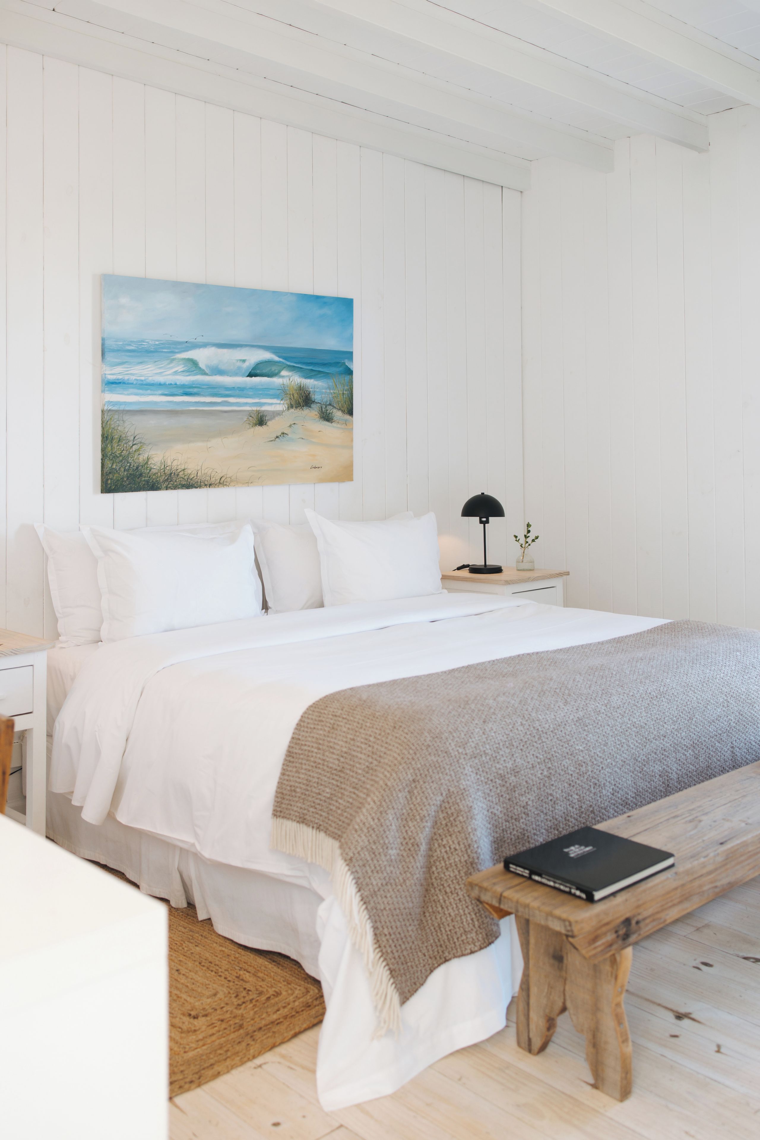 A bedroom with a bed, a bedside table with a lamp, a bench, and a painting of a beach scene.