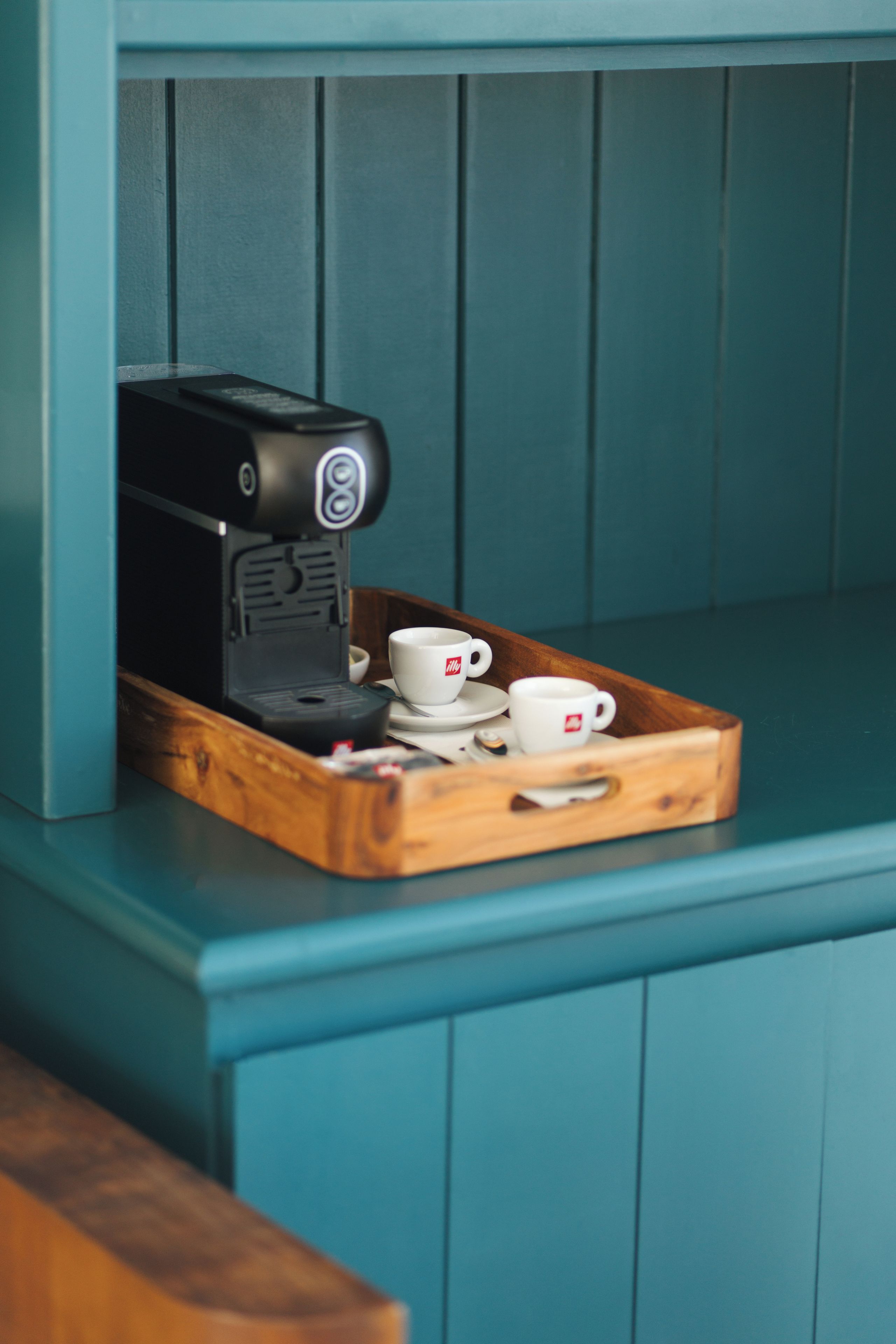 Una máquina de café sobre una bandeja de madera con dos tazas en un banco azul verdoso.
