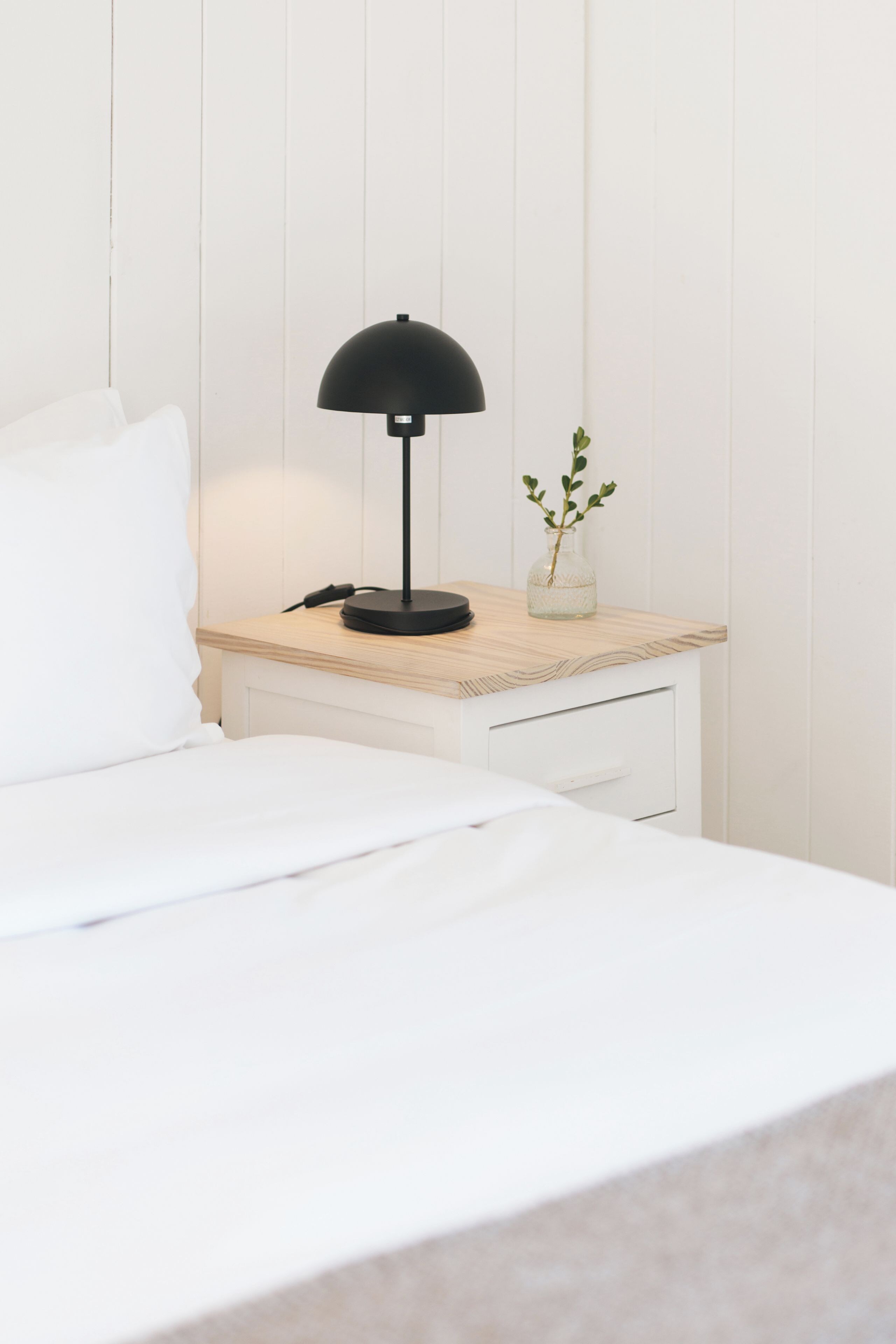 A neatly made bed with white bedding, a small bedside table with a black lamp, and a vase with a plant.