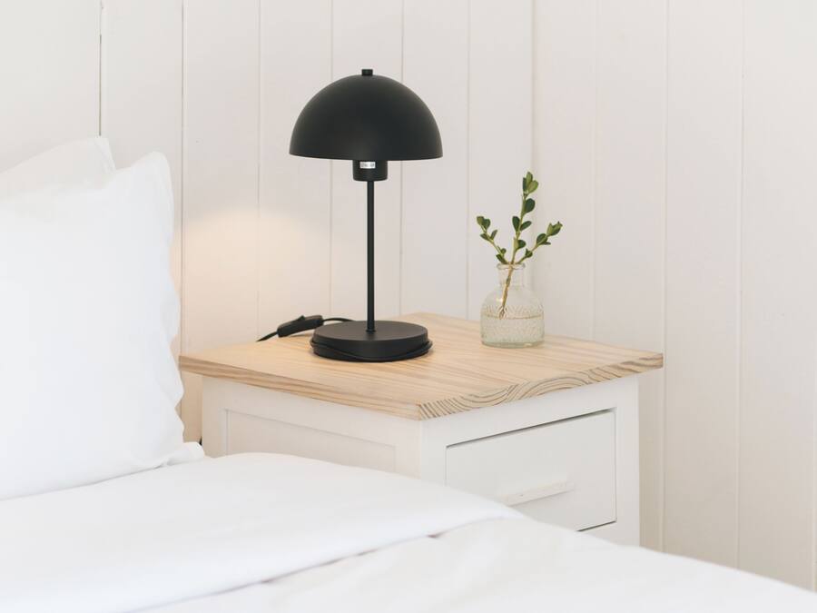 A neatly made bed with white bedding, a small bedside table with a black lamp, and a vase with a plant.