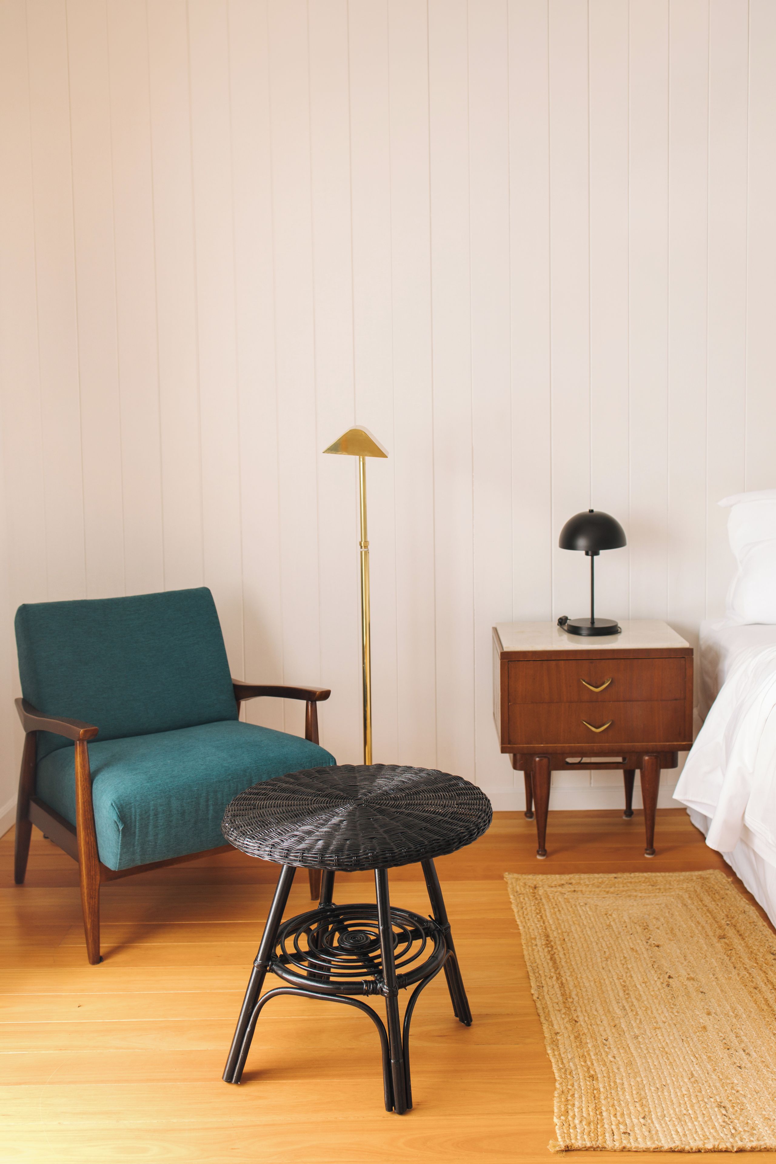 A room with a teal armchair, a black wicker stool, a floor lamp, a bedside table with a lamp, and a bed with white bedding.