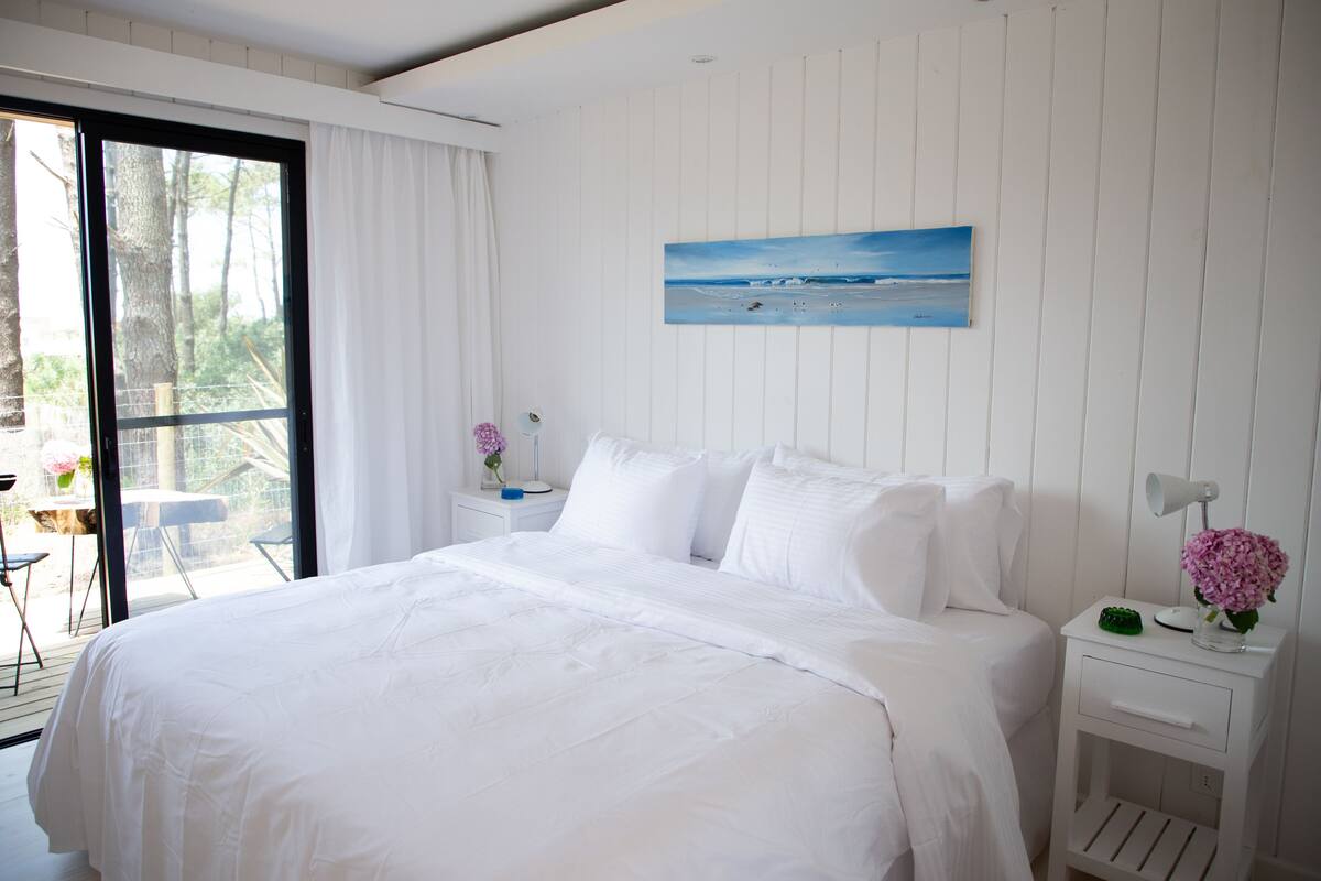 A bedroom with a large bed, a nightstand with a lamp, a painting of a beach, and a view of the outdoors through a sliding door.