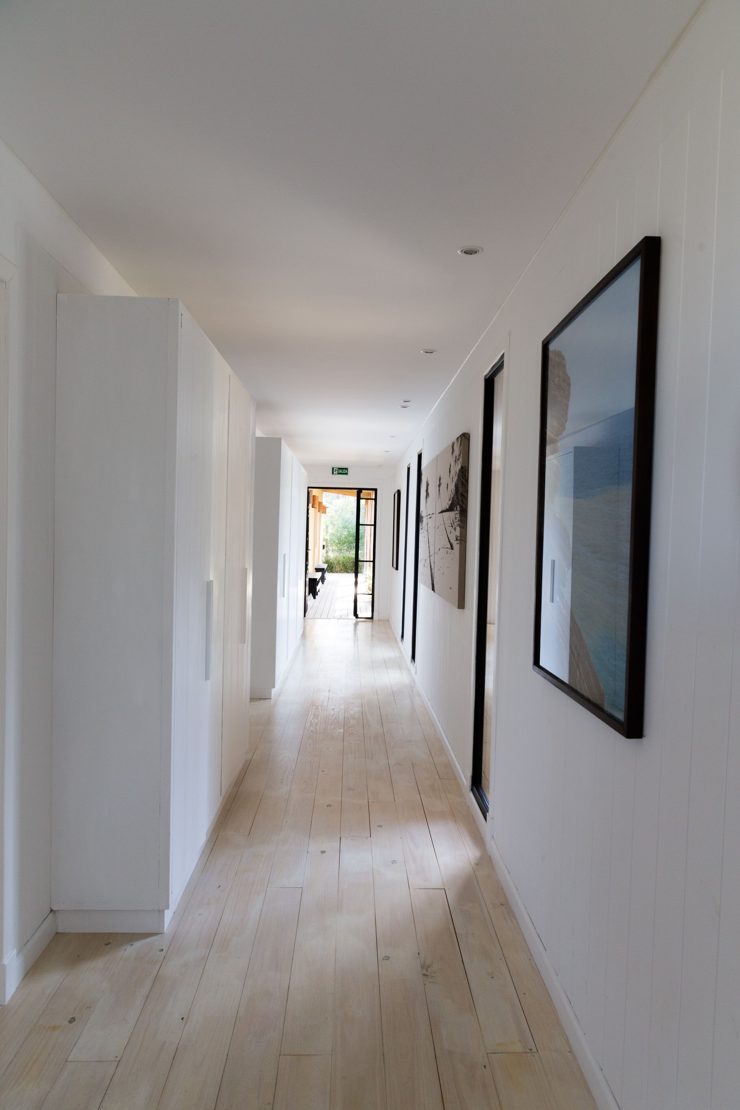 A long, narrow hallway with light wooden floors and white walls. There are several framed artworks on the walls, and a door at the end of the hallway leads to an outdoor area with a bench and plants.