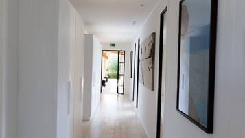 A long, narrow hallway with light wooden floors and white walls. There are several framed artworks on the walls, and a door at the end of the hallway leads to an outdoor area with a bench and plants.