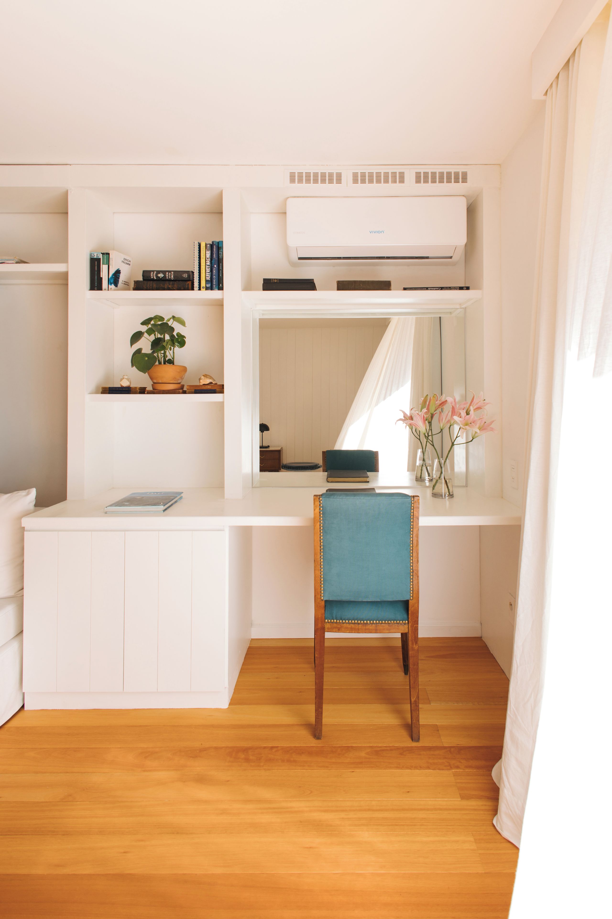 Ein kleines Zimmer mit weißem Schreibtisch, einem Stuhl, einem Blumentopf und einem Fenster mit Vorhängen.