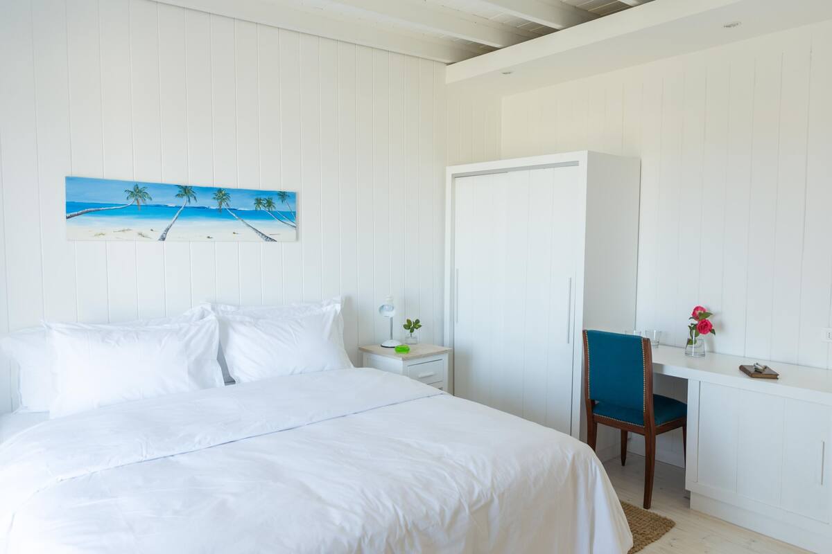 A bedroom with a bed, a desk, a chair, a wardrobe, and a painting of a beach scene.