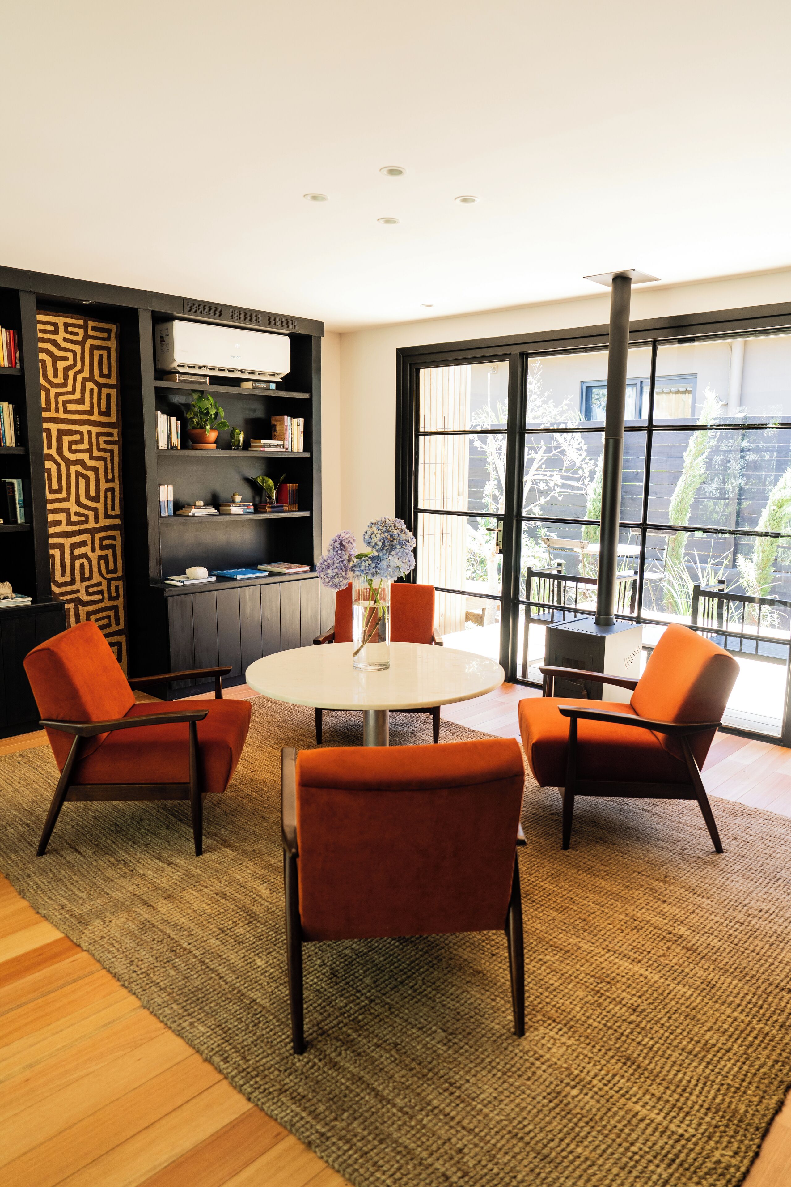 A modern living room with a round table, orange chairs, a bookshelf, and a large window with a view of the outdoors.