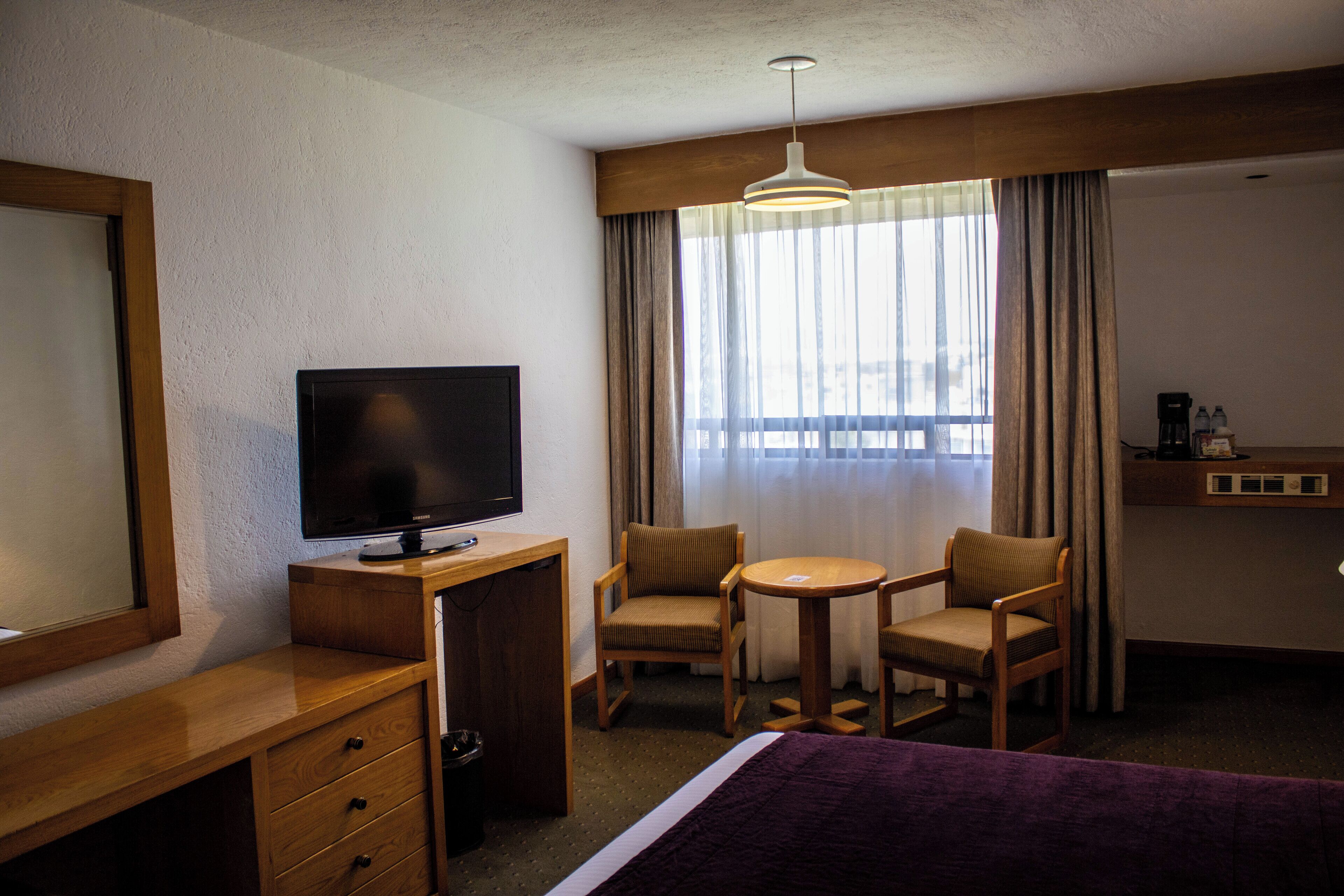 A hotel room with a bed, a desk with a TV, two chairs, a small table, and a window with curtains.