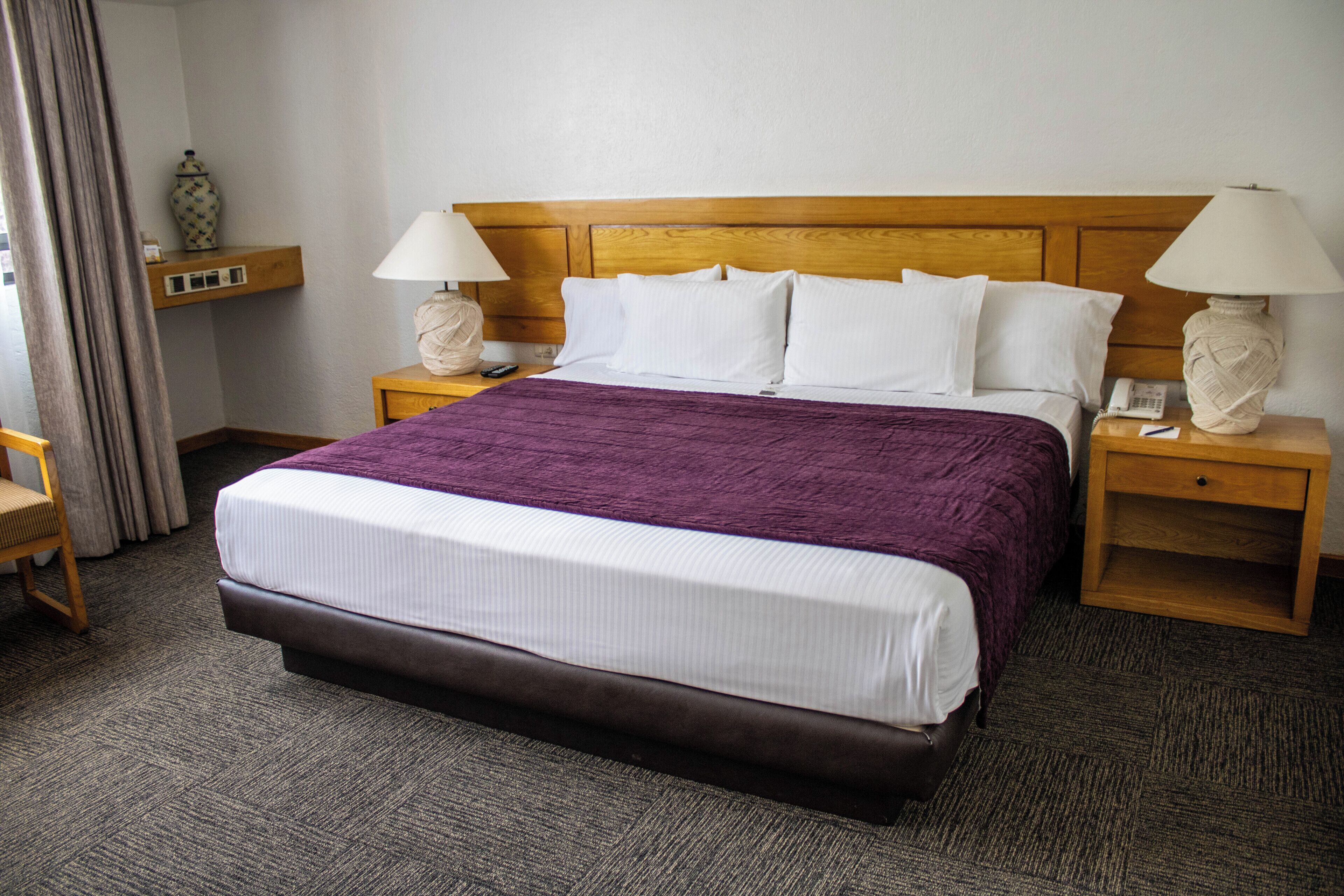A hotel room with a large bed, two bedside tables, a wooden headboard, and a small shelf with a vase.