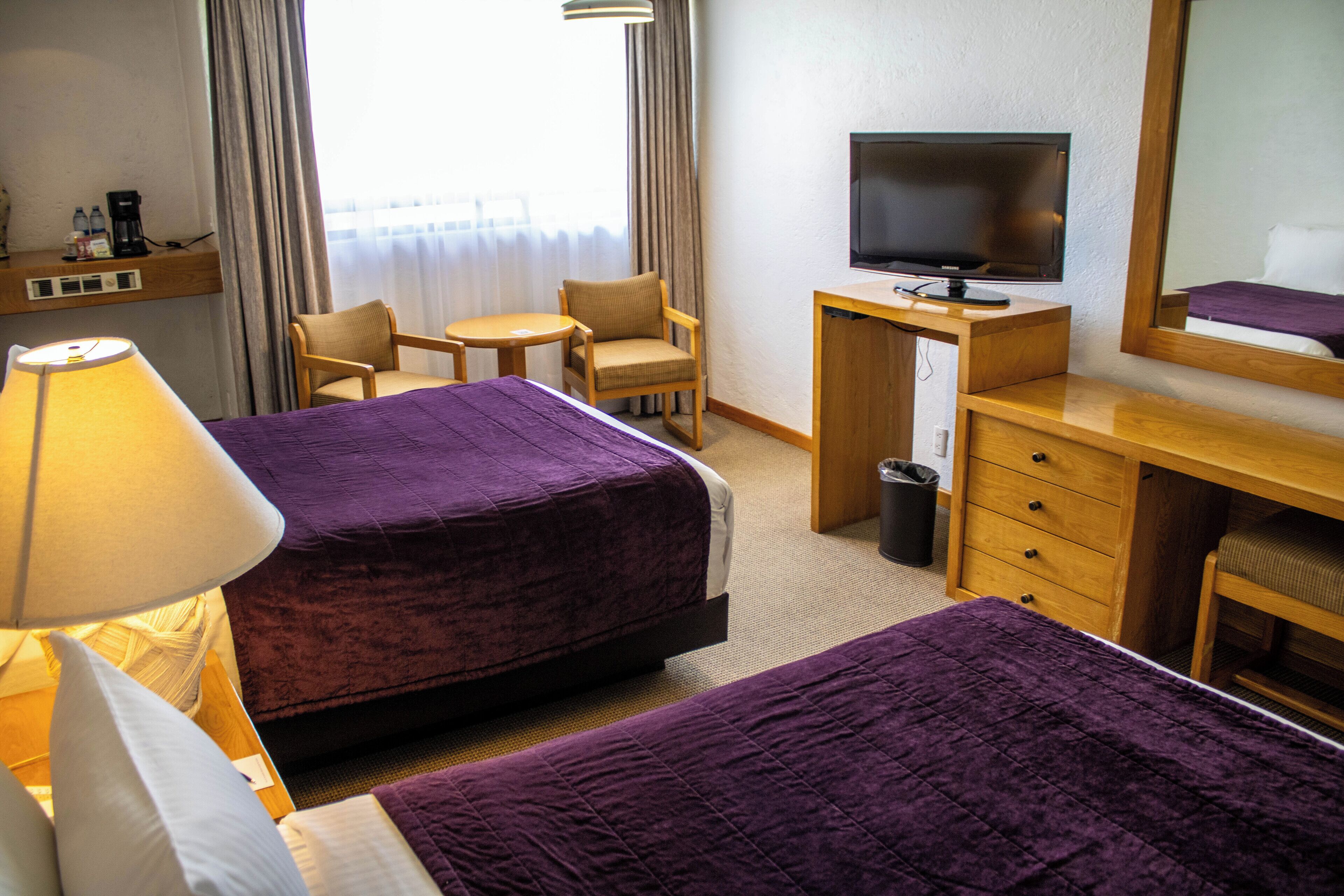A hotel room with two beds, a desk with a TV, a chair, a lamp, and a window with curtains.
